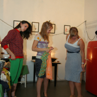 Three women are in a room decorated with framed pictures and string lights, clearly in preparty mode. One woman in a red sweater and green pants looks at her phone. The second woman, wearing a polka dot top, holds an orange-and-green fabric. The third woman in a striped dress looks down at her feet. A red appliance is seen on the right side at Maiņas punkts.