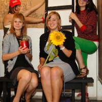 Four women are posing for a photo indoors. Two are seated on a bench, one holding a red apple and the other a sunflower. The other two women are standing behind them, with one wearing a red hat and the other in sunglasses and green pants. A ladder and abstract painting in the background add to the preparty vibe.