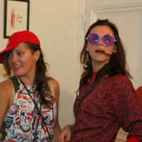 Two women having fun indoors, likely at a preparty. One wears a red cap, white tank top with a pattern, and large earrings. The other sports large purple sunglasses, red earrings, and a red patterned top. Both are smiling and enjoying themselves. A framed picture and a white door are in the background.