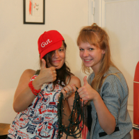 Two young women are standing indoors, smiling and giving thumbs up. The one on the left is wearing a red cap, a sleeveless top with various prints, and holding tangled string lights. The one on the right has blonde hair, wears a gray blazer, and is also giving a thumbs up. It looks like they're ready for their preparty at Maiņas punkts!