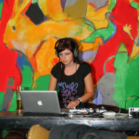 A DJ with headphones is focused on her laptop while performing at the preparty. She is wearing a black shirt with purple text and is surrounded by colorful, abstract wall art in the background. On the table, there are various DJ equipment pieces, including cables and a bottle. A jacket is draped over a chair in the foreground.