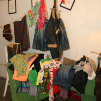 A cluttered display features various clothes hanging and piled on a table covered with a green tablecloth, resembling an eclectic preparty vibe. Garments include shirts, jackets, skirts, and pants in diverse colors and patterns. Above, strings of fairy lights and framed sketches can be seen on the wall. A red cap hangs on a rack.