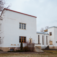 A two-story, Mazirbe-style white rectangular building with flat roofs and minimal architectural details features large windows and a small staircase leading to a door on the right. The exterior shows weathering and cracks, while sparse landscaping surrounds it under an overcast sky.