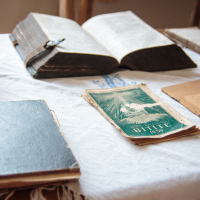 A table draped in white cloth displays several old books, with an open tome at the center, framed by closed volumes with worn covers. A vintage 1937 booklet titled "The Bible" adds to the scene's nostalgic warmth. This cozy setting evokes memories of a quiet Mazirbe evening.