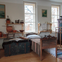 A vintage bedroom in Mazirbe features a single wooden bed adorned with a patterned blanket. Shelves showcase pottery and rustic items. A window reveals bare trees outside, while an armoire stands beside a spinning wheel. The walls display landscape paintings, enhancing the nostalgic atmosphere.