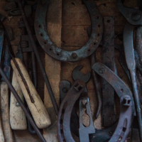 A wooden drawer, possibly reminiscent of Mazirbe's old-world charm, is filled with various blacksmith tools, including rusty horseshoes, pliers, wrenches, and tools with wooden handles. Aged and darkened by frequent use, the items are set against a rustic backdrop that enhances the vintage feel.