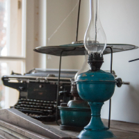 A vintage scene unfolds in Mazirbe, highlighting an old-fashioned typewriter and a blue oil lamp with a clear glass chimney atop a wooden chest. In the background, a balance scale by the window filters soft, natural light, evoking nostalgia and charm.