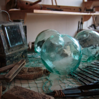 A rustic workbench in Mazirbe displays antique fishing gear: three large glass fishing floats, a weathered tin lantern, wooden net weights, and fishing nets. Assorted tools, including scissors, are scattered around, with shelves in the background holding more vintage items.