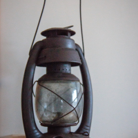 A vintage black metal lantern, reminiscent of Mazirbe's coastal charm, rests gracefully on a light wooden surface. The lantern boasts a glass globe and a crisscross metal frame. Its sturdy handle arches above for ease of carrying, set against a plain off-white wall.