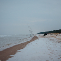 A solitary figure sits near fishing rods planted in the sand along a tranquil, overcast beach in Mazirbe. The shoreline curves gently, and the ocean waves lap softly onto the snow-dusted beach. A dense line of evergreen trees is visible in the distance against the cloudy sky.