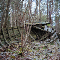A decaying wooden boat is nestled among tall trees in a forest near Mazirbe. The boat, covered in moss and surrounded by fallen leaves and branches, rests on a forest ground lightly dusted with snow. The trees form a dense backdrop, hinting at a cold, serene winter setting.