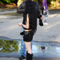 A person with long black hair, dressed in black shorts, a black top, and tall black boots, stands facing another person in a brown jacket and blue jeans. The scene resembles a moment from a 2009 metalshow. In the background, another person walks amid greenery while a puddle glistens on the pavement near their feet.