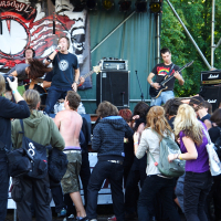 A crowd at an outdoor metalshow enthusiastically watches a rock band perform on stage. The lead singer, dressed in a black shirt, is energetically engaging with the audience. The guitarist in a colorful shirt plays near an amplifier. Some of the attendees, their dyed hair reminiscent of 2009 trends, appear excited and energetic. There is a band banner in the background and trees are visible.