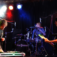 Three musicians perform on stage under colorful lights. The focus is on a guitarist wearing a dark shirt on the left, adjusting equipment at this 2009 metalshow. In the background, a drummer plays a drum set, and on the right, another guitarist in a light shirt faces the first guitarist. A dark backdrop completes the scene.
