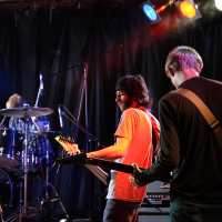 Three musicians are performing on stage. The drummer is playing a drum set, while two guitarists stand in front, one facing away and the other with long hair wearing glasses, looking down at his instrument. Colorful stage lights overhead illuminate the scene at Metalshow '09. The backdrop is black.