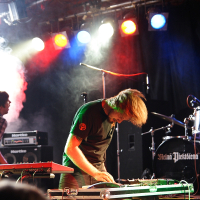 Two musicians perform energetically on stage at a 2009 metalshow, bathed in vibrant colored lights. One, wearing a black shirt, leans over an electric keyboard, hair swinging. The other, in a plaid shirt, is partially obscured. Smoke and stage equipment, including drums and amplifiers, fill the background.