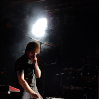 A person with medium-length hair performs on stage at a 2009 metal show, illuminated by a bright spotlight creating a smoky effect. They lean over a keyboard or synthesizer and hold a microphone close to their mouth. In the background, drumming equipment can be seen with "Mek Megtetem" written on the bass drum.