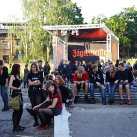 A group of people gather outside at a Jägermeister event. Some sit on steps in front of a stage with a Jägermeister banner, reminiscent of the Metalshow '09 atmosphere, while others stand or sit on the ground nearby. They appear to be socializing, and many are dressed in casual, dark-colored clothing. Trees and buildings are in the background.