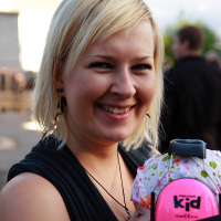 A smiling woman with short blonde hair is outdoors holding a baby, both enjoying what seems to be a lively Metalshow. The baby wears a white hat with colorful patterns and bright pink ear protection headphones labeled "kid '09." The woman, in black clothing and earrings, has blurred people and buildings in the background.
