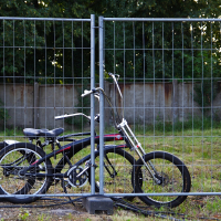 A black bicycle with a long front fork is locked to a wire mesh fence using a U-lock. The bike, reminiscent of the rides seen at Metalshow 2009, is parked on a grassy area with some exposed soil and scattered vegetation. Behind the fence, there is a concrete wall and a backdrop of trees in a wooded area.