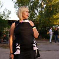 A woman with short blonde hair stands outside on a paved area with trees in the background, reminiscent of a scene from 2009. She wears a black dress and holds a baby in a black wrap carrier, their legs clad in black and white striped leggings and pink shoes protruding. Other people reminiscent of a metalshow are in the background.