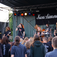 A band performs on an outdoor stage with a banner behind them reading "Sanctimony." The lead vocalist sings into a microphone, while other members play electric guitars and drums. The audience below, comprised of people with long hair and back-printed shirts, watches the 2009 metalshow.