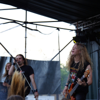 Two musicians performing on an outdoor stage at Metalshow '09. The one on the left is singing into a microphone, while the one on the right, with long blonde hair, is playing a bass guitar. Both are wearing black T-shirts, and the stage backdrop is dark with a partially visible drum kit. An audience with long hair is visible in the foreground.