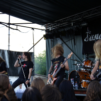 Band performing on outdoor stage at Metalshow '09. Five male members with long hair are playing instruments including guitars, bass, and drums. Lead singer is gripping a microphone stand. Audience with blond and brown hair visible in foreground. Backdrop features the word "Sanctuary.