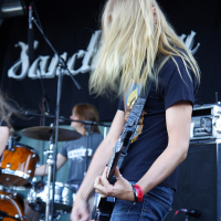 A person with long blonde hair is playing a guitar on stage at Metalshow '09, moving their head vigorously. They are wearing a black t-shirt and jeans. In the background, another person is playing drums. The backdrop features the partially visible word "Sanctuary" in stylized, white lettering.