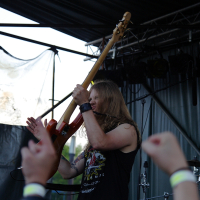 A long-haired musician in a black sleeveless shirt with a colorful design plays an orange electric guitar on an outdoor stage at Metalshow '09. He is captured mid-performance raising his arm. The audience, with raised arms and wristbands, is visible in the foreground. Stage lights and equipment are in the background.