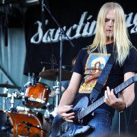 A long-haired musician plays an electric guitar on an outdoor stage at a 2009 metalshow. He wears a black T-shirt with a colorful design. Behind him are a drum set and other musical equipment. A black banner with the word "Sanctuary" is hung in the background.