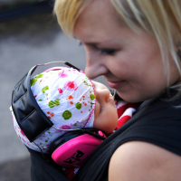 A woman with blonde hair is carrying a baby with floral-patterned headwear in a baby carrier at Metalshow '09. The baby, wearing pink ear protection headphones, appears to be asleep. The woman is smiling and looking down fondly at the baby while the background is out of focus.