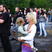 A woman with blonde hair, sunglasses, and a colorful skirt holds a young child in her arms at Metalshow 2009. The child wears a striped shirt and green vest. They are surrounded by other attendees, some in dark clothing, with greenery in the background. Several people observe something off-frame.