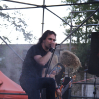 A lead singer on stage passionately sings into a microphone while a guitarist with long hair headbangs in the background at this 2009 metalshow. The stage features metal scaffolding and a red canopy. Trees and some blurred audience members are visible in the background, adding to the outdoor concert ambiance.