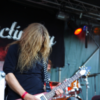 At a 2009 Metalshow, a musician with long blond hair covering their face plays an electric guitar on stage. They are wearing a black shirt and have a red wristband. The guitar has a distinct V shape with a white body and dark edges. Behind them, a drummer plays under red and orange stage lights.