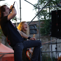 Two musicians perform passionately on an outdoor stage at the 2009 metalshow event. The lead singer, with long hair and a black t-shirt, leans forward while gripping the microphone. The bassist, also with long hair, plays intensely beside him. Fans' hands and stage equipment are visible, with a brick building in the background.