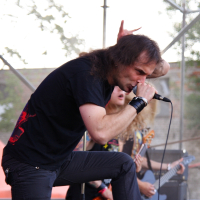 A man wearing black clothing, including a black shirt and pants, is performing on stage at a 2009 Metalshow. He grips a microphone with his right hand, his left fist raised. A musician with long blonde hair and a bass guitar stands in the background. Metal scaffolding or stage equipment is visible around them.