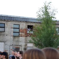 A crowd of people is gathered in an outdoor area near a worn-out two-story brick building. Several attendees of Metalshow '09 look out of a large window on the building's second floor. The building shows signs of decay with patches of missing bricks, while a tall tree stands in front.