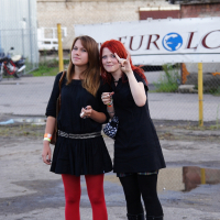 Two young women stand outdoors in front of a chain-link fence with a EuroLC sign, reminiscent of an industrial metalshow setting. One, with red hair and a black dress, makes a peace sign while holding ice cream. The other, with brown hair in a black dress and red tights, looks off to the side. It's 2009; bikes and industrial structures complete the background.