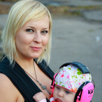A blonde woman with a slight smile carries a baby in a black baby carrier. The baby is wearing a white hat with colorful patterns, pink earmuffs labeled "kid," and a striped sweater. They are outdoors at an event, possibly reminiscent of the bustling streets during Metalshow 2009, with a blurred urban background.