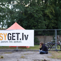 A person sits on the ground outside, leaning against a metal fence. Behind the fence, a large white banner reads "EASYGET.lv pasākumi, jaunumi, foto, apskati." Nearby are two bicycles and an orange tent set up in front of dense green trees. It's clear this spot is ready for Metalshow '09.