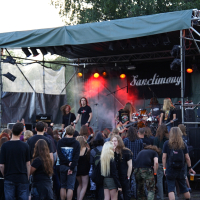 A crowd gathers in front of an outdoor stage where a metal band is performing at Metalshow '09. The band members are engaged with their instruments and singing under colorful stage lights. The audience, dressed in dark clothing, watches attentively. Trees and outdoor equipment are visible in the background.