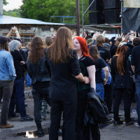 A crowd of people, mostly dressed in black, gathers outdoors for a Metalshow '09 concert. In the foreground, a person with long brown hair and a black coat faces a person with red hair in a black outfit. Stage equipment and speakers are visible in the background, and the ground is wet with puddles. Trees and buildings are also visible.
