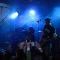 At a 2009 metalshow, a rock band performs on a dimly lit stage with blue and purple lights and smoke effects. The vocalist, standing at the center, sings into a microphone while the bassist on the right plays his instrument. Fans in the foreground raise their hands, showing their excitement and appreciation.