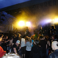 A dynamic 2009 metalshow scene at night with a rock band performing on stage under vibrant yellow and orange lights. The energetic crowd in the foreground includes fans dancing, headbanging, and raising their hands. Stage equipment and musicians are partially visible through the stage smoke.