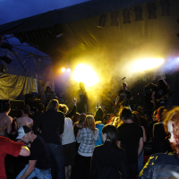 A crowd of people at an outdoor metalshow stands closely together and faces a stage where a band is performing under bright yellow and white stage lights. The atmosphere is lively, with some concertgoers visibly dancing. The sky is dark, indicating it is nighttime, reminiscent of the energetic gigs of 2009.