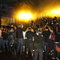 A crowd of people at an outdoor rock concert at night faces a lit-up stage. The stage, illuminated by bright yellow lights, showcases musicians performing intensely. Audience members in dark clothing headbang and raise their hands in unison. The muddy, wet ground only adds to the raw energy of Metalshow '09.