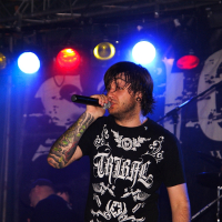 A man on stage singing into a microphone at a 2009 metalshow. He has shoulder-length dark hair, tattooed arms, and wears a black T-shirt with white ornate designs and text. In the background, colorful stage lights in red, yellow, and blue illuminate the scene, and a drummer is partially visible.