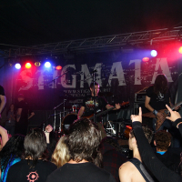 A rock band performs on a dimly lit stage with colored lights, featuring a backdrop with "STIGMATA '09" in bold letters. The lead singer holds a microphone, and the guitarist, bassist, and drummer are engaged in their instruments. The energetic crowd at this metalshow raises their hands, some forming rock-on signs.
