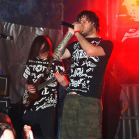 A vocalist passionately sings into a microphone while a guitarist plays beside him on stage during Metalshow '09. Both are dressed in black graphic t-shirts. The dim-lit venue is illuminated by vibrant red stage lights, and the audience, raising hands, is visible in the foreground.